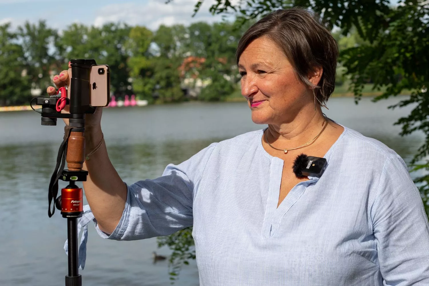Frau steht an einem See, vor ihr steht ein Stativ bei dem oben ein Smartphone mit einem speziellen Halter montiert ist. Die Frau tippt auf dem Handydisplay. An der Bluse der Frau ist ein Ansteckmikrofon befestig. 