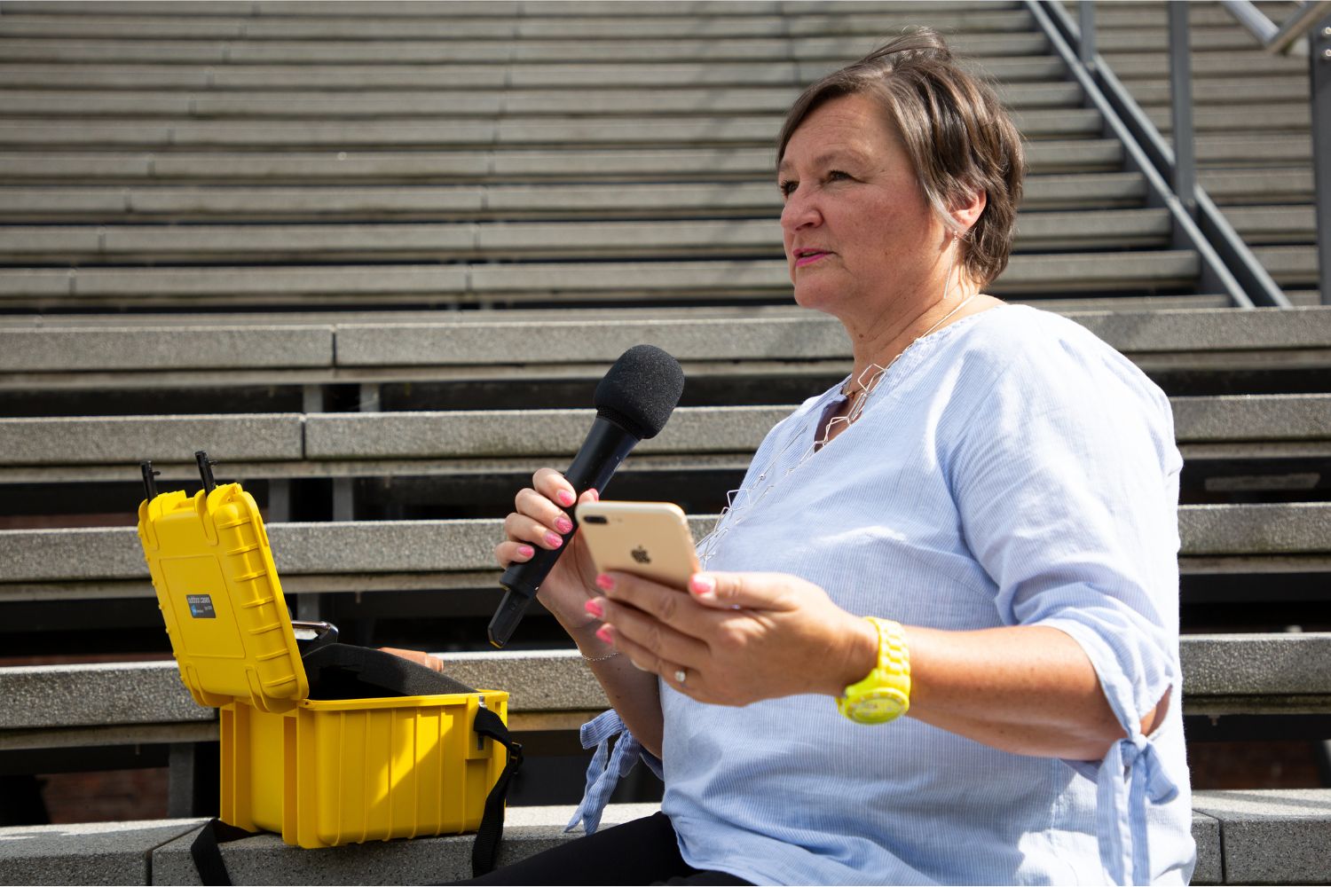 Heike sitzt auf der Treppe in der einen Hand ein Mikrofon, in der anderen ein iPhone. Sie schaut in die Ferne, während sie in das Mikrofon spricht. Vor ihr auf der Treppe steht ein kleiner gelber Equipent-Koffer.